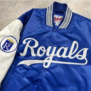 May include: A royal blue satin varsity jacket featuring white sleeves and the word Royals embroidered in white script across the chest. The jacket has a striped ribbed collar, snap button closures, and a Kansas City Royals logo patch on the sleeve.