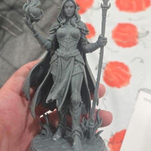 Peut inclure: Figurine grise d'un personnage fantastique féminin, tenant une sphère enflammée et un bâton. La figurine porte une armure et une cape flottante, debout sur une base circulaire. La figurine mesure environ 15 cm de haut.