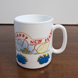 Puede incluir: Taza de cerámica blanca con asa, con las palabras "HAPPY NEW YEAR!" en letras rojas. La taza está decorada con diseños florales azules y amarillos y detalles dorados. Un artículo festivo y decorativo.