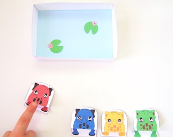 May include: Four paper frog cutouts with different colored bodies and black eyes. The frogs are sitting on a white surface in front of a blue paper box with two green lily pads and pink flowers.