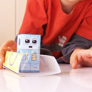 May include: A blue and white paper robot toy with a yellow and white paper boat. The robot has a face with two large eyes and a small mouth. The boat has a handle on the top.