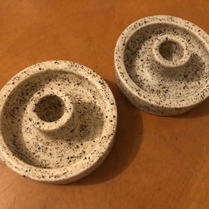 May include: Two speckled ceramic candle holders. The candle holders are circular with a central hole and a raised outer rim. The speckled pattern features black spots on a cream-colored background. The candle holders are on a wooden surface.