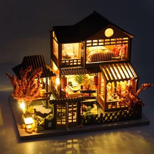 Puede incluir: Una casa japonesa en miniatura con iluminación interior cálida. La estructura de madera presenta múltiples niveles, incluyendo un dormitorio y un comedor, con un pequeño jardín y linternas decorativas. El diseño general es acogedor y atractivo.