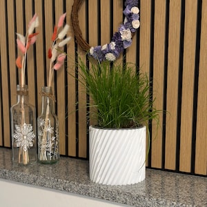 Puede incluir: Una exhibición decorativa en estantería con una maceta blanca con diseño en espiral con hierba verde, dos botellas de vidrio con detalles florales y una corona de flores moradas y blancas. El fondo es una pared de listones de madera.