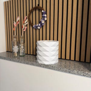 Puede incluir: Una maceta geométrica blanca con una superficie texturizada se encuentra sobre una repisa de granito. Dos botellas de vidrio con diseños florales y flores secas están a la izquierda. Una pequeña corona cuelga de una pared de listones de madera.