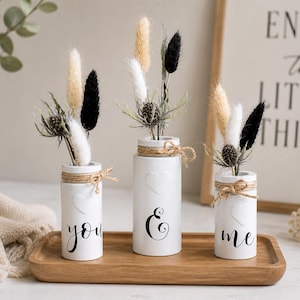 May include: Three white ceramic vases with dried floral arrangements, each with a jute twine accent. The vases are inscribed with the words "you", "&", and "me" in black script. The vases are on a wooden tray.