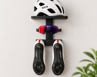May include: A black wall-mounted storage rack holding a white KASK bicycle helmet on the top shelf, a pair of Oakley sunglasses with red and blue lenses in the center, and a pair of white DMT cycling shoes with black soles hanging below.