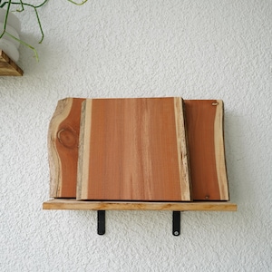 May include: A wooden shelf with three rectangular planks of reddish-brown wood. The shelf is supported by two black metal brackets and is mounted on a textured white wall. The wood has a natural, unfinished appearance.