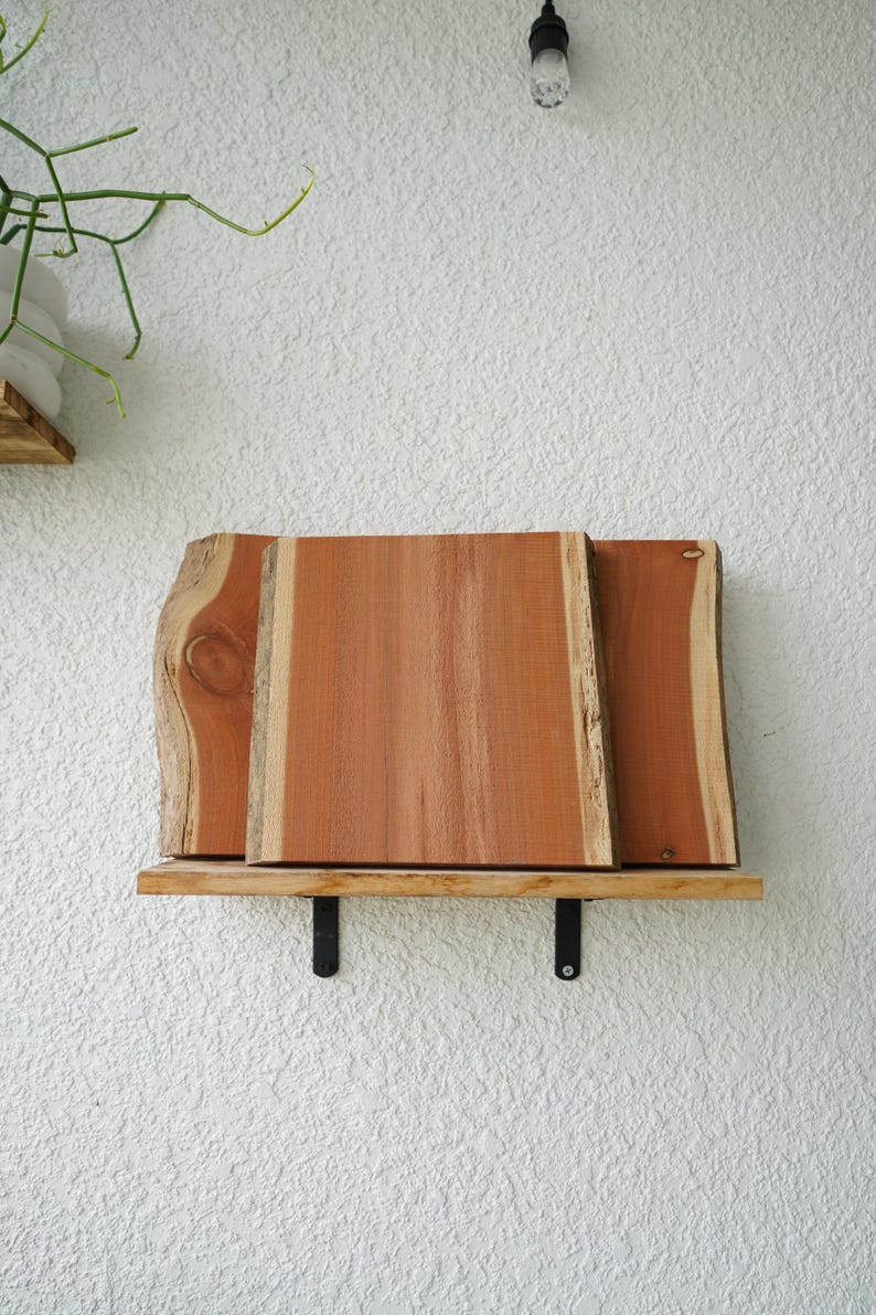 May include: A wooden shelf with three rectangular planks of reddish-brown wood. The shelf is supported by two black metal brackets and is mounted on a textured white wall. The wood has a natural, unfinished appearance.