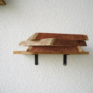 May include: A wooden shelf with three stacked, irregularly shaped wooden planks. The shelf is supported by black metal brackets against a textured white wall. The wood has a reddish-brown hue.
