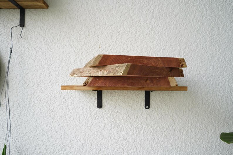 May include: A wooden shelf with three stacked, irregularly shaped wooden planks. The shelf is supported by black metal brackets against a textured white wall. The wood has a reddish-brown hue.