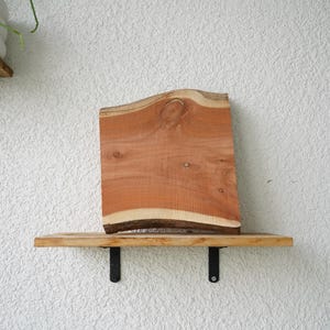 May include: A wooden shelf with a live edge, displaying a reddish-brown wood slab. The shelf is supported by black metal brackets against a textured white wall. The wood slab has a natural, organic shape. The shelf is approximately 60 cm long.
