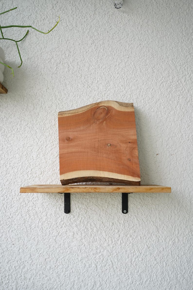 May include: A wooden shelf with a live edge, displaying a reddish-brown wood slab. The shelf is supported by black metal brackets against a textured white wall. The wood slab has a natural, organic shape. The shelf is approximately 60 cm long.
