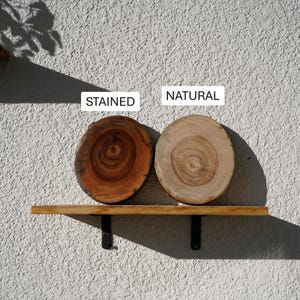K&ouml;nnte beinhalten: Zwei Holzscheiben, eine gebeizt und eine nat&uuml;rlich, auf einem Holzregal mit schwarzen Metallhalterungen. Die gebeizte Scheibe ist ein sattes Braun, w&auml;hrend die nat&uuml;rliche Scheibe hellbraun ist. Die W&ouml;rter "STAINED" und "NATURAL" sind aufgedruckt.