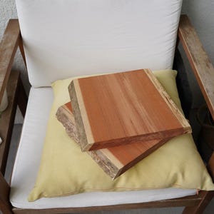 May include: Two rectangular, unfinished wooden slabs with natural edges, stacked on a yellow cushion. The wood is reddish-brown with lighter streaks. The cushion rests on a white cushion of a wooden chair.