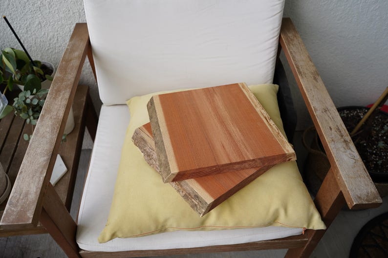 May include: Two rectangular, unfinished wooden slabs with natural edges, stacked on a yellow cushion. The wood is reddish-brown with lighter streaks. The cushion rests on a white cushion of a wooden chair.