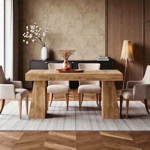 May include: A light-colored wooden dining table with a rectangular top and two thick, angled legs. Four beige upholstered chairs surround the table, set on a gray rug. A vase with flowers and a copper vase with dried plants are on the table.