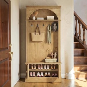 May include: A tall light oak wood hall tree featuring an arched top shelf, three metal hooks holding a woven tote bag and a scarf, a hanging hat, and a lower storage bench with two open shelves containing multiple pairs of shoes and umbrellas.