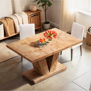 May include: A rectangular wooden dining table with a light brown finish. The table has a unique pedestal base and is surrounded by two white chairs. A vase of red flowers and a bowl of fruit decorate the table.