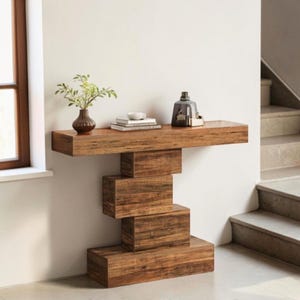 May include: A wooden console table with a unique, geometric design. The table features a long, rectangular top supported by stacked, rectangular blocks. A small vase with greenery, books, and decorative items are on the top. The wood has a warm, natural finish.
