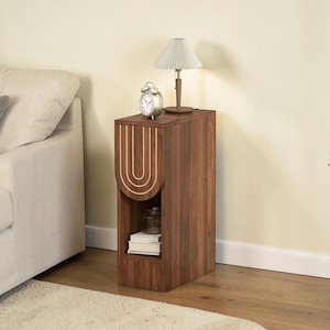 May include: A dark brown wooden side table with a unique arched design on the front. The table features a small open shelf and is topped with a small lamp and a silver alarm clock. The table is next to a beige sofa.