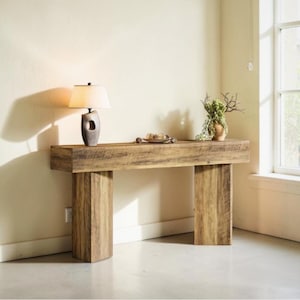May include: A rustic wooden console table with a rectangular top and two thick, vertical supports. A lamp with a beige shade and a decorative vase with greenery sit on the table. The table is made of light brown wood.