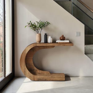 May include: A wooden console table with a unique, curved design. The table has a natural wood finish and a shelf holding decorative vases and books. A vase with green foliage adds a touch of nature. The table is placed against a neutral-colored wall.