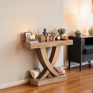 May include: A light oak console table with a unique crossed-leg design. The table is adorned with decorative items, including books, candles, figurines, and a small clock. A pillow with a Union Jack design rests on the lower shelf. The table is placed against a neutral wall.