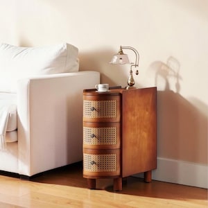 May include: A narrow wooden bedside table with three rounded drawers featuring woven cane fronts and small dark knobs. A small white ceramic cup and a brass-toned desk lamp with a glass shade sit on the flat top surface of the cabinet.