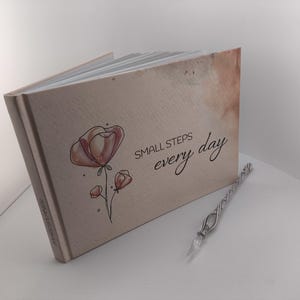 Könnte beinhalten: Ein Tagebuch mit den Worten "SMALL STEPS every day" und einer floralen Illustration. Das Tagebuch ist geöffnet und zeigt leere Seiten. Ein dekorativer Glasstift liegt neben dem Tagebuch.
