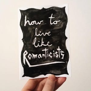 Puede incluir: Una tarjeta blanca con el texto manuscrito "how to live like Romanticists" en blanco sobre una forma negra irregular. La tarjeta está sujeta con una mano.