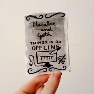Peut inclure: Une carte dessinée à la main avec les mots "Macabre and Goth" et "Things to do Offline" à l'encre noire. La carte présente un fond de lavis gris et des volutes décoratives.