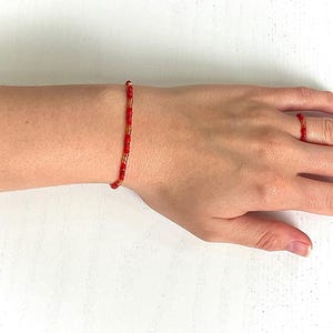Könnte beinhalten: Ein rotes und goldenes Perlenarmband- und Ringset. Das Armband befindet sich an einem Handgelenk und der Ring an einem Finger. Die Perlen sind klein und zylindrisch, mit einer Mischung aus roten und goldenen Farben. Der Schmuck ist zart und scheint handgefertigt zu sein.