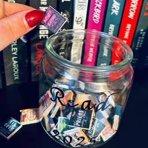 Pode incluir: Um frasco de vidro transparente cheio de miniaturas de livros, com a palavra "Read 2023" escrita no frasco. Uma miniatura de livro é segurada acima do frasco. Livros são visíveis ao fundo.