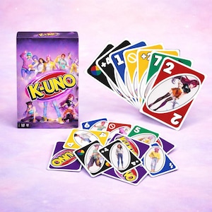 May include: A K-UNO card game set featuring a purple box with the game's logo and character illustrations. The cards are fanned out, displaying various colors and numbers, with character art on some. The game is set against a soft, pink background.
