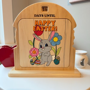 May include: Wooden Easter countdown calendar featuring a bunny, flowers, and Easter eggs. The calendar reads "26 Days Until Happy Easter!" in colourful lettering. The frame is light brown with a wooden base.