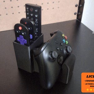 Puede incluir: Organizador negro para mandos a distancia y mando de juegos. El organizador tiene capacidad para tres mandos a distancia y un mando de juegos. Los mandos a distancia son negros con botones de varios colores. El mando de juegos es negro con botones de colores.
