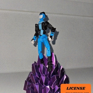 Puede incluir: Una figura de acción azul y negra se alza sobre una formación de cristales morados. La figura tiene articulaciones y está en posición de pie. La base es un cúmulo de formas geométricas. La imagen incluye el texto "LICENSE - COMMERCIAL USE - AUTHOR ATTRIBUTION REQUIRED".