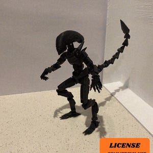 Puede incluir: Una figura de acción articulada de Xenomorfo negro con cola segmentada y cabeza texturizada. La figura está posada sobre una superficie de color claro. Un cuadrado naranja con texto está en la esquina inferior derecha.
