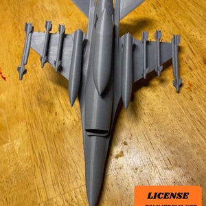 May include: A gray 3D-printed model of a fighter jet, complete with wings, tail, and multiple missiles attached. The model is resting on a wooden surface. An orange square with text that says "LICENSE - COMMERCIAL USE - AUTHOR ATTRIBUTION REQUIRED" is in the bottom right corner.