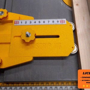 May include: A yellow wood strip jig with a ruler marked in centimeters. The jig has a slot and is secured with star-shaped knobs. The text "STRIP JIG" is visible. The jig is positioned next to a piece of wood.