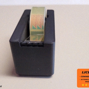 Puede incluir: Un dispensador de cinta adhesiva negro con un rollo de cinta adhesiva en su interior. El rollo de cinta adhesiva tiene la marca "TESA" impresa. El dispensador está sobre una superficie clara.