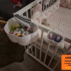 Könnte beinhalten: Weißes Babybett mit wolkenförmigen Aufbewahrungskörben, die Babyspielzeug und Bücher enthalten. Ein kleinerer wolkenförmiger Behälter enthält Schnuller. Das Bett hat ein rosa gemustertes Laken und eine geflochtene Bettumrandung.