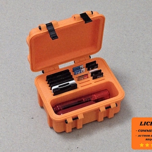 May include: An open orange storage case with black latches and hinges. Inside are various small black and white items, and a red pen. The case is on a gray surface. A small orange square with text is in the lower right corner.