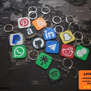 Puede incluir: Un conjunto de llaveros cuadrados con logotipos de redes sociales y tecnología, como Pinterest, Amazon y LinkedIn. Cada llavero tiene un anillo y una cadena plateados. El fondo es un mapa del mundo.