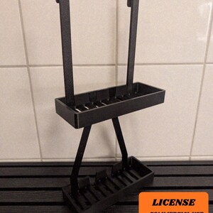 May include: A black metal shower caddy with two rectangular trays and two vertical bars with curved tops. The caddy is designed to hang over a shower head. The caddy is shown against a white tile background.