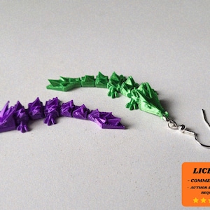 May include: A pair of dragon-shaped earrings in green and purple. The earrings are made of a 3D-printed material and feature a detailed, textured design. One earring is attached to a silver-coloured ear wire. The image also includes a license notice.