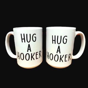May include: Two white ceramic mugs with the text "HUG A HOOKER" in black, bold letters. The mugs have a curved handle and a light tan base. The background is solid black.