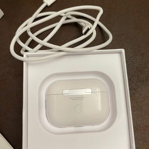 May include: A white charging case for wireless earbuds, nestled in a white box, with a white charging cable. The case has a smooth, rounded design with a silver accent. The text "Designed by Apple in California Assembled in China" is printed on the case.