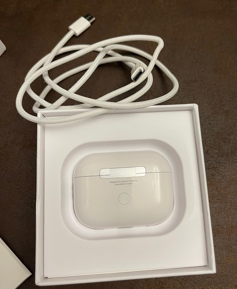May include: A white charging case for wireless earbuds, nestled in a white box, with a white charging cable. The case has a smooth, rounded design with a silver accent. The text "Designed by Apple in California Assembled in China" is printed on the case.